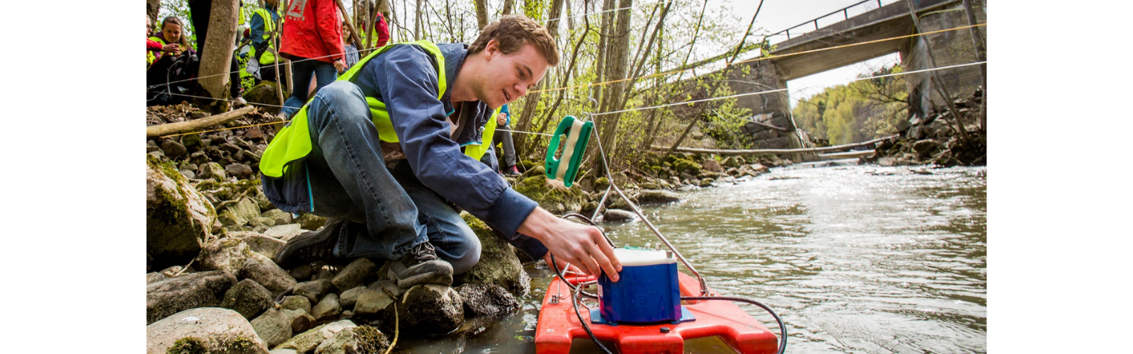 A person wearing a high-vis yellow vest is kneeling on rocks to place a blue box on a floating red raft on a flowing river. 